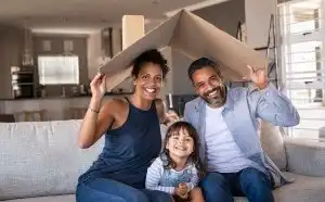 Two parents hold a cardboard roof over their child. They are all smiling.